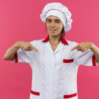 pleased young female cook wearing chef uniform points to herself on isolated pink background with copy space